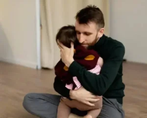 A man sitting on the floor, cradling a baby in his arms.