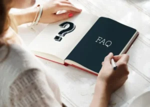 A woman holding a pen, sitting at a table with a notebook that has "FAQ" written on it.