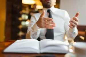 Man sitting at a table with a book in front of him, gesturing as if explaining something
