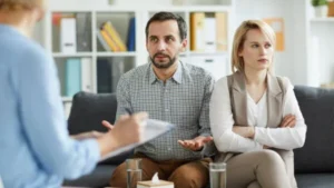 A couple conversing on a couch, in front of a person holding a pen and paper.