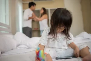 A young girl sitting on a bed between her mother and father having a discussion in the background.