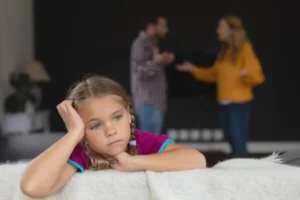 A young girl sitting on a couch with her parents in the background.