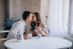 A couple sharing a tender kiss at a table, with a child sitting nearby.