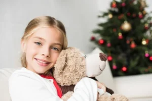 A young girl holding a teddy bear in front of a Christmas tree.