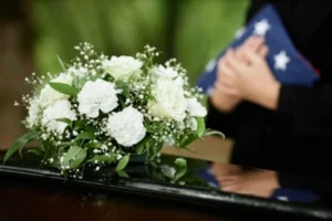 A casket with a white flower bouquet and a person holding an American flag beside it.