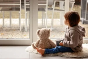 A baby is looking out the glass door while holding a mug, with a teddy bear beside him.