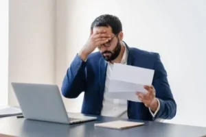 A professional man in a suit sits at his desk, focused on a piece of paper and a laptop.