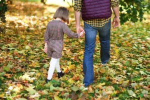 A father holds his daughter's hand as they walk through fallen leaves.