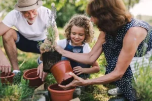 Two adults and a child plant flowers outdoors; the child holds a plant while one adult assists.