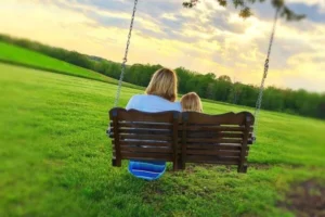 A mother and daughter sit on a wooden swing chair, surrounded by green grass and trees.