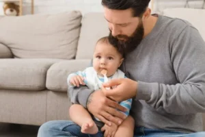 A father is sitting on the floor while feeding his baby.