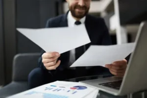 A man in a suit holding two papers with a laptop on the table.