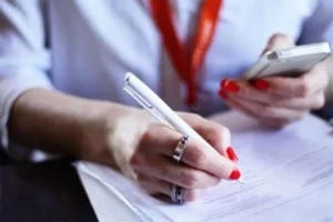 A woman with red nail polish holding a pen and a phone, with paper underneath.
