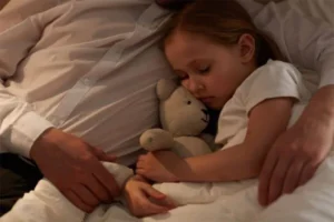 A little girl hugging a teddy bear while her father wraps his arms around her.