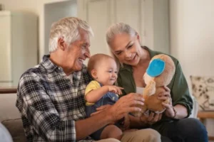 The grandfather carries his grandchild while the grandmother gives a teddy bear to the baby boy.