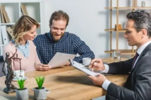 A couple sitting at a table in an office, with a man in a suit writing on a piece of paper.