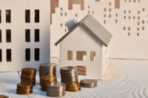A white wooden house model with a stack of coins in front.