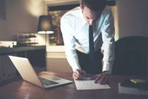 A businessman stands while writing on a piece of paper next to a laptop on the table.