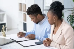 A husband is signing a document as his wife watches, waiting for him to complete his signature.