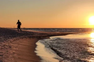 A person runs along a beach at sunset, with ocean waves nearby and the sun setting on the horizon.