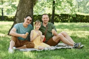 Two men sitting on the grass with a young lady between them, enjoying a casual moment together