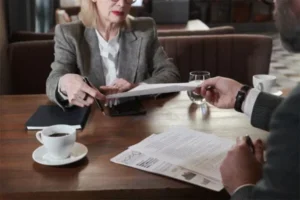 Man gives a document to a woman holding a pen, with coffee, a notebook, and water on the table.