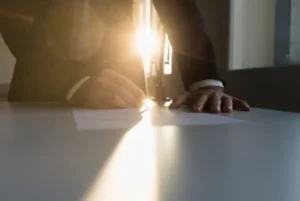 A man in a suit signs a document while leaning on a table, illuminated by sunset rays.